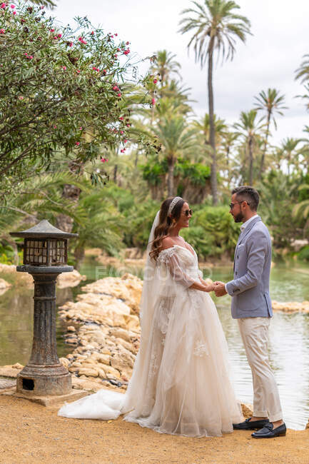 Side view full length of happy young married couple in wedding clothes standing and holding hands while looking at each other near lake and green trees in daytime in garden — Stock Photo