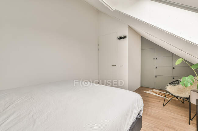 Letto con lenzuolo bianco situato vicino alla parete bianca nella moderna camera da letto soffitta luce — Foto stock