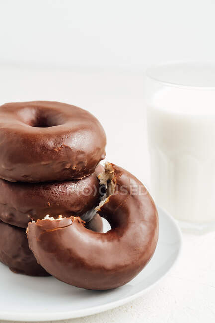 Pilha de doces saborosos donuts com esmalte de chocolate colocado na placa na mesa branca com vidro de leite na sala de luz — Fotografia de Stock