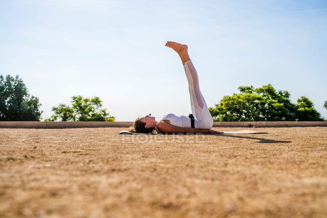 Vista laterale di una donna calma in activewear bianco che esegue Halasana su un tappeto a terra nel parco contro alberi verdi e cielo blu senza nuvole nella giornata di sole — Foto stock