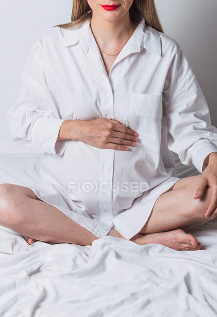 Вид спереду анонімних молодих вагітних ніжних жінок, які торкаються животик, сидячи на ліжку — стокове фото