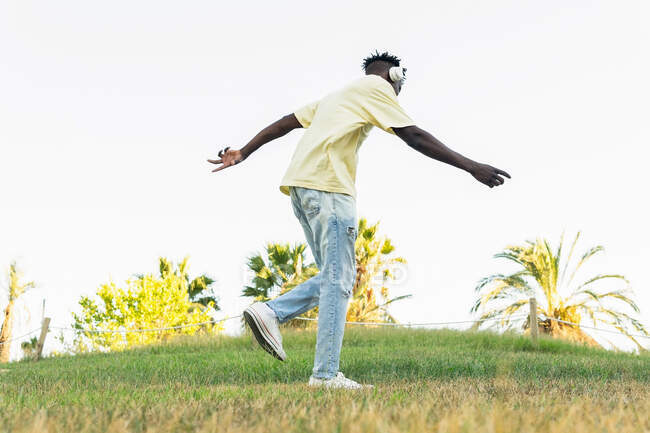 Снизу сзади вид неузнаваемого афроамериканского молодого мужчины в повседневной одежде, слушающего музыку с наушниками в солнечный день в парке — стоковое фото