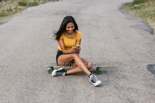 Full body of positive ethnic female browsing on smartphone while sitting longboard on asphalt road — Stock Photo