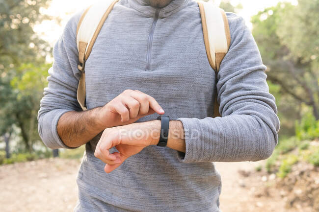 Crop unrecognizable male hiker in casual clothes and backpack using smartwatches while walking along narrow path in green forest — Stock Photo