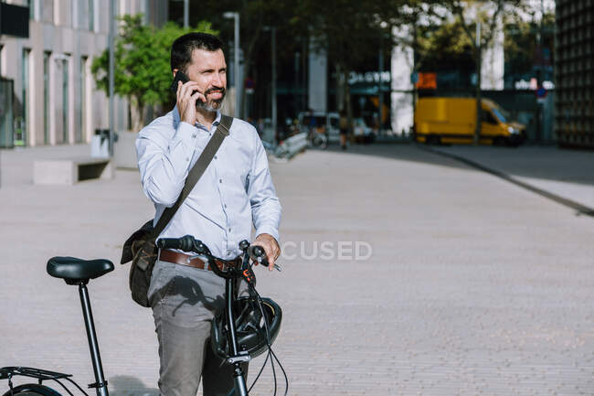 Travailleur masculin en tenue formelle debout avec vélo et avoir une conversation sur téléphone portable au centre-ville — Photo de stock
