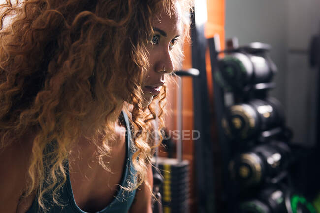 Vista lateral de la deportista concentrada de cultivo con pelo largo y rizado mirando hacia adelante contra pesas en el gimnasio durante el día - foto de stock