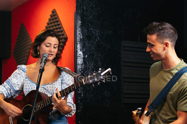 Jovem positivo tocando guitarra perto de músico feminino tocando guitarra acústica e cantando em clube de luz — Fotografia de Stock