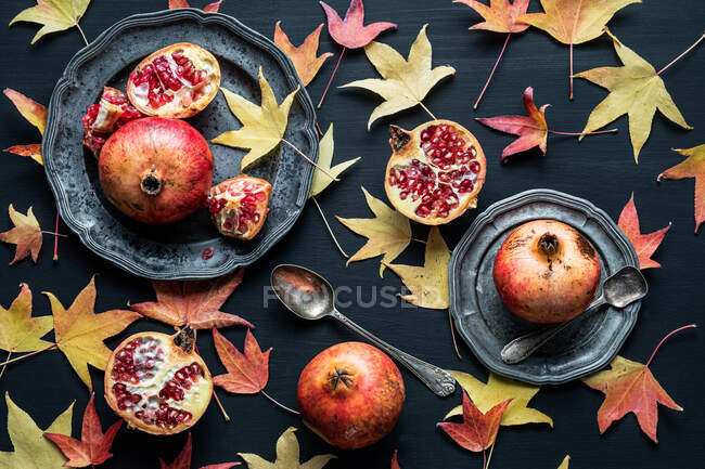 Top view composition of dried autumn leaves placed near plates of ripe sweet pomegranates on black background — Stock Photo