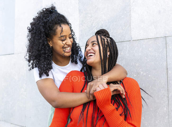 Feliz namoradas multiétnicas em roupas da moda de pé e abraçando contra a parede cinza na rua da cidade durante o dia — Fotografia de Stock