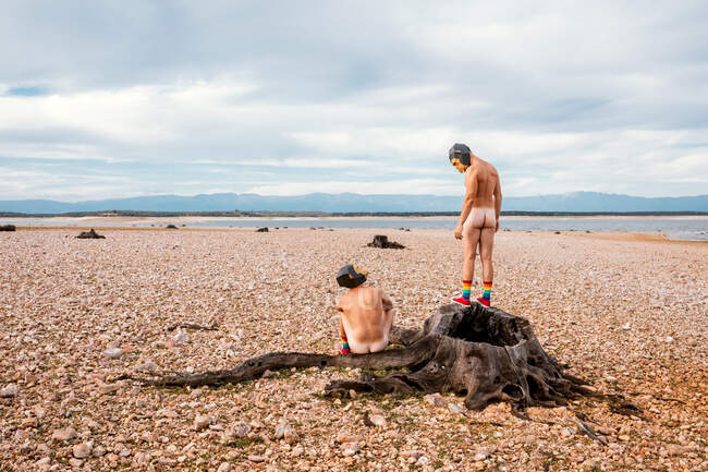 Naked men in monkey masks looking at camera while spending time on stone seashore in sunny day — Stock Photo