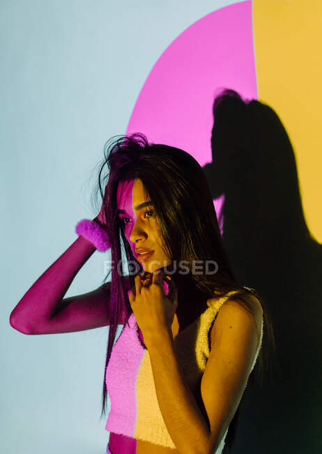Trendy junge hispanische Frau mit erhobenem Arm sieht weg gegen Schatten und bunte Projektorlichter auf grauem Hintergrund — Stockfoto