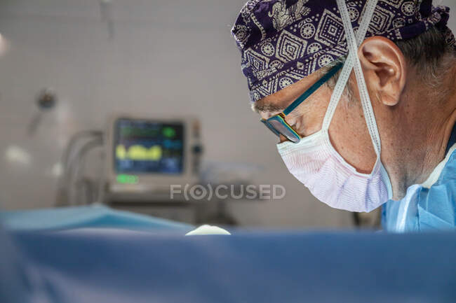 Vecchio chirurgo maschio in maschera protettiva e uniforme con cappello che lavora in sala operatoria durante l'intervento — Foto stock
