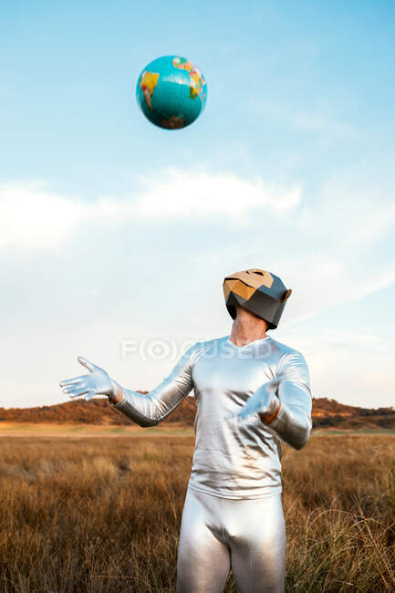 Man in geometric monkey mask and silver latex suit tossing Earth painted ball in yellow grassy field in sunny day — Stock Photo