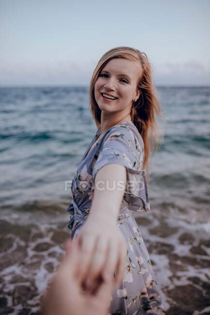 Sorrindo jovem fêmea em vestido casual em pé na praia de areia perto do mar ondulante, enquanto segurando a mão da colheita pessoa irreconhecível no verão — Fotografia de Stock