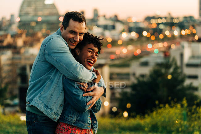 Romántica pareja diversa abrazando y mirando hacia otro lado mientras está de pie en el césped contra el paisaje urbano con edificios sobre fondo borroso - foto de stock