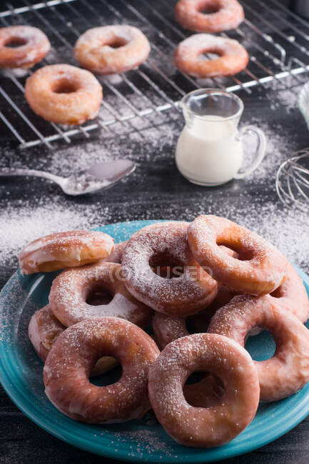 Sweet fried doughnuts served on plate near metal cooling rack and jug of milk on black messy table with powdered sugar — Stock Photo