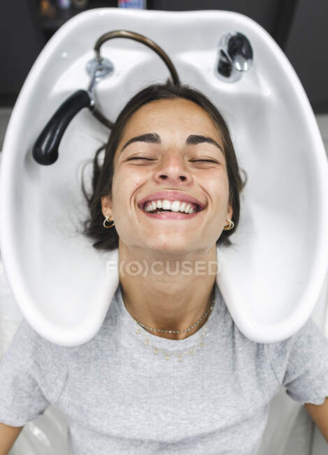 Vista superior de la joven dama positiva en ropa casual con los ojos cerrados mientras se sienta cerca del fregadero contragolpe mientras se prepara para lavar el cabello en el salón de peluquería ligera - foto de stock