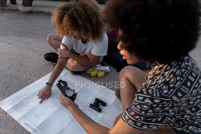 Alto angolo di escursioniste afroamericane concentrate sedute con gambe incrociate e percorso di pianificazione con mappa e bussola — Foto stock