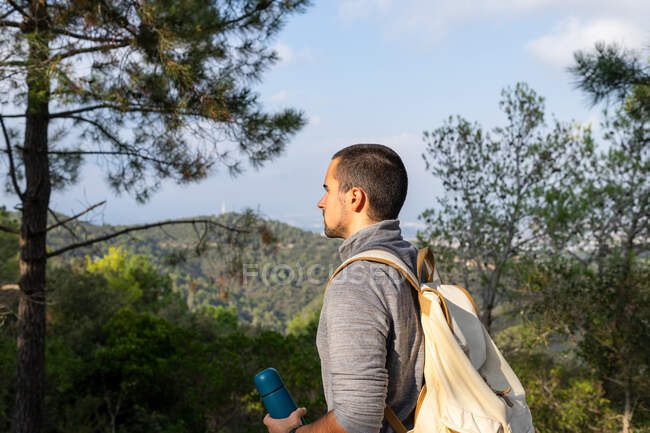 Pensivo jovem barbudo hispânico caminhante masculino em roupas casuais de pé em exuberante floresta verde com termo na mão e olhando para longe no dia ensolarado — Fotografia de Stock
