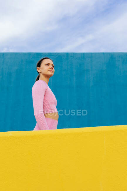 Visão lateral da fêmea concentrada grave com cabelo escuro vestindo roupas ativas rosa claras olhando para longe contra a parede azul — Fotografia de Stock