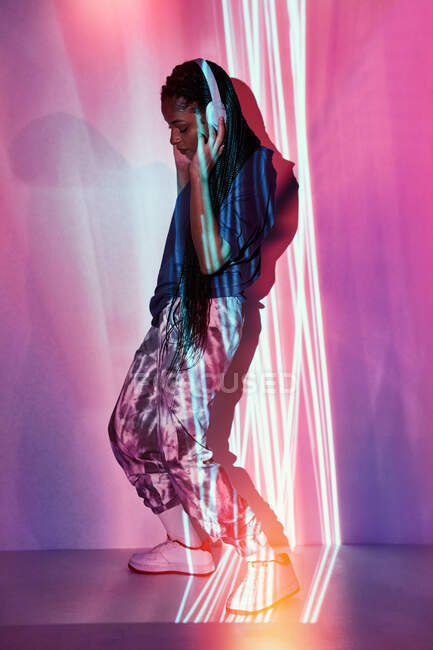Vista lateral de un adolescente étnico de moda en un elegante atuendo bailando mientras escucha música en auriculares parados cerca de la pared con iluminación brillante - foto de stock