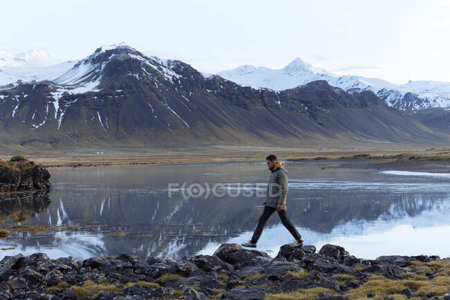 Vista lateral del joven viajero masculino en ropa de abrigo caminando a lo largo de la costa pedregosa del lago tranquilo rodeado de montañas nevadas durante el viaje en Islandia - foto de stock