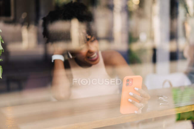 Durch das Fenster einer fröhlichen jungen ethnischen Frau mit dunklem Afro-Haar im trendigen Outfit und echten drahtlosen Kopfhörern, die glücklich lächelt, während sie Videotelefonie auf dem Smartphone in einem modernen Café führt — Stockfoto