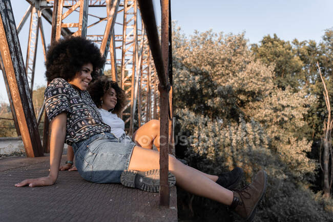 Seitenansicht einer glücklichen afroamerikanischen Mutter, die in der Nähe ihrer Tochter lächelt, die bei Sonnenuntergang auf einer verwitterten Brücke sitzt — Stockfoto