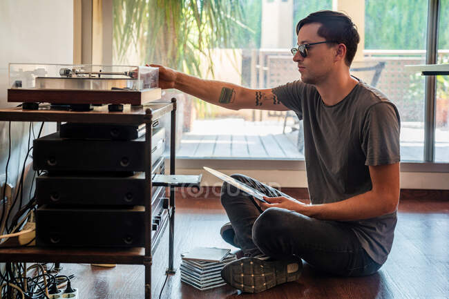 Side view of male in casual outfit sitting on floor near turntable and looking at vinyl record in hand — Stock Photo