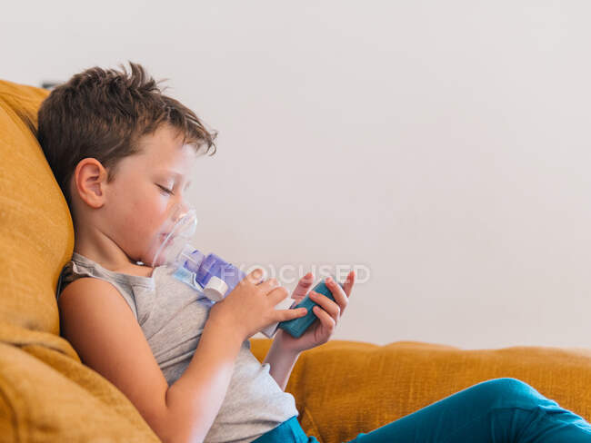 Задоволений хворий хлопчик використовує дихальний пристрій і вдихає кисень від інгалятора, сидячи вдома на дивані. — стокове фото
