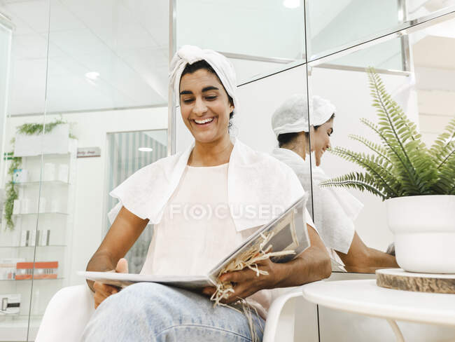 Веселая этническая клиентка с полотенцем на голове сидит у зеркала и смотрит альбом в современном салоне — стоковое фото