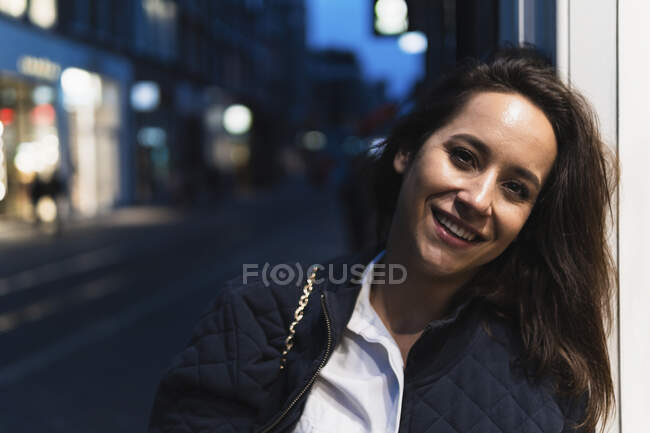 Happy female in jacket standing on city street near contemporary buildings and empty road while smiling and looking at camera — Stock Photo
