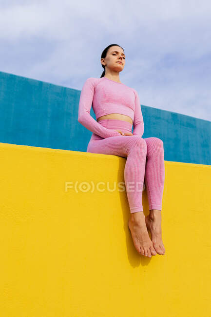 Low angle full body of confident fit female in pink sportswear sitting with closed eyes on bright yellow surface — Stock Photo