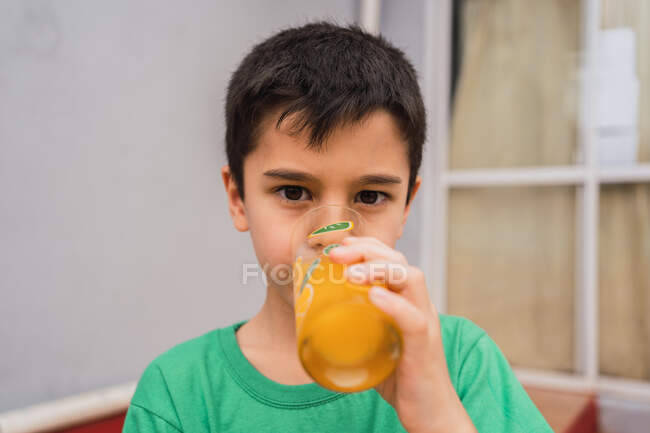 Серйозний хлопчик у сорочці з чорним волоссям дивиться на камеру, п'ючи напій зі скла в світлій кімнаті вдома — стокове фото