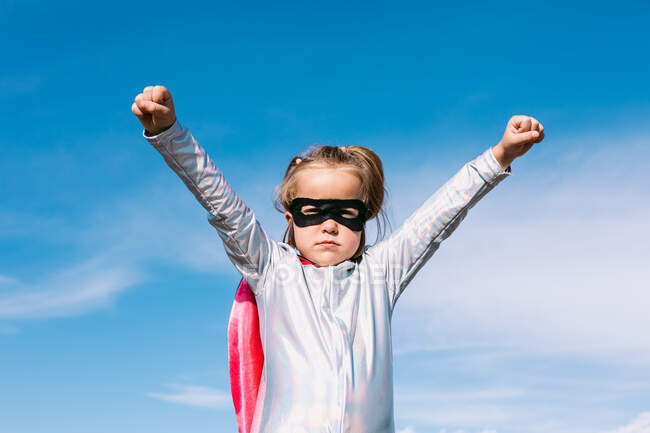 From below small girl in superhero costume raising outstretched fists for showing power while standing against blue clear sky — Stock Photo
