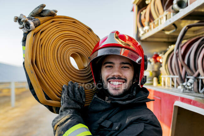 Tapferer männlicher Feuerwehrmann mit Schutzhelm und Uniform blickt in die Kamera, während er in der Nähe eines Feuerwehrfahrzeugs auf einem Acker einen schweren Schlauch auf der Schulter trägt — Stockfoto
