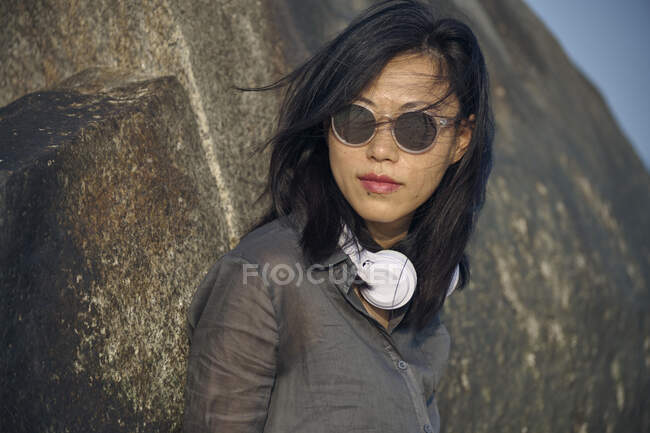 Mujer asiática tranquila con pelo negro volador en gafas de sol de moda y auriculares blancos modernos de pie cerca de la formación rocosa en la naturaleza - foto de stock
