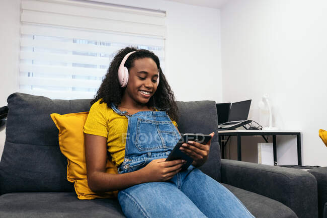 Снизу счастливая афроамериканка в стильном наряде слушает музыку в наушниках и пользуется планшетом, сидя на сером диване в светлом помещении дома — стоковое фото