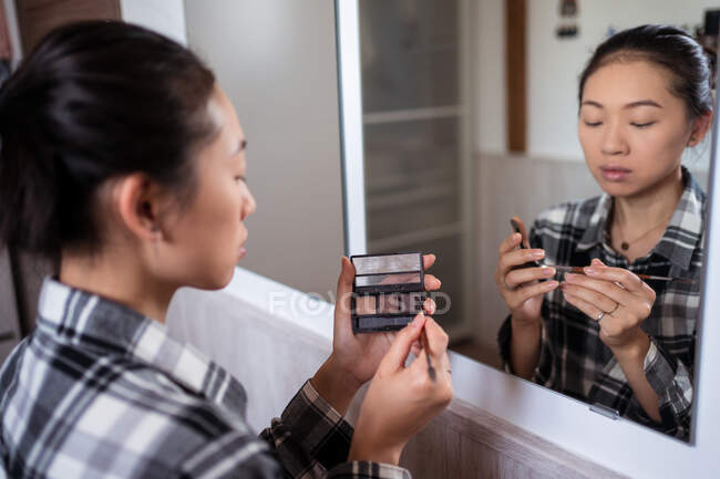 Focused charming ethnic female doing eyebrow with brush and palette while doing makeup and looking in mirror — Stock Photo