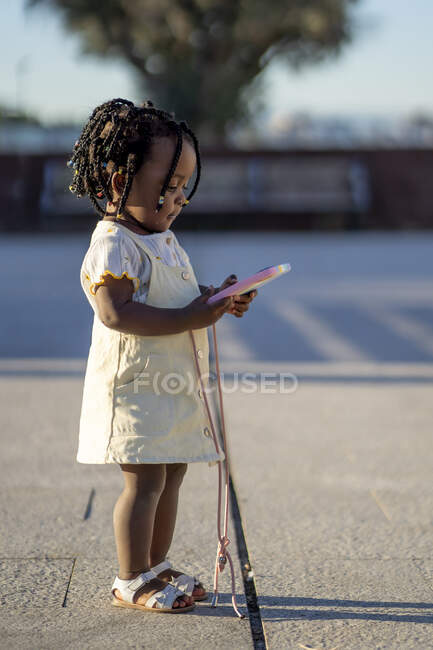 Вид сбоку на спокойную маленькую афроамериканскую девочку с черными косичками в стильной одежде, стоящую со смартфоном в красочном футляре в руках на асфальтовой дорожке на улице в солнечный день — стоковое фото