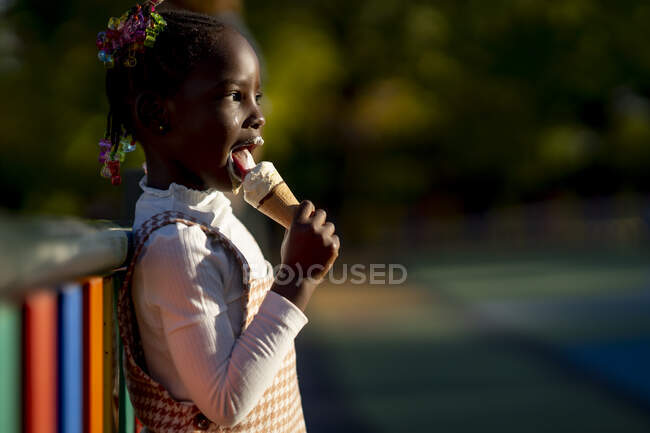 Vista lateral de la alegre chica afroamericana de pie cerca de la valla de colores y lamiendo helado dulce en la calle contra el fondo borroso - foto de stock