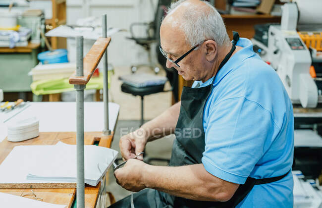 Vista lateral do artesão masculino sênior atencioso em avental e óculos amarrando fitas em placa de madeira antes de trabalhar na máquina de impressão — Fotografia de Stock