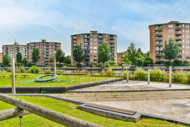 View of empty park with benches and green lawn and trees located near modern residential buildings in city on sunny summer day in bright daylight — Stock Photo