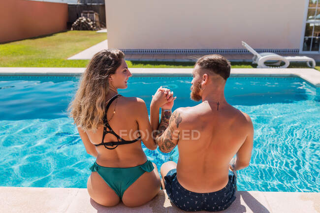 Вид ззаду любляча пара в купальниках дивиться один на одного і тримає руки, доглядаючи за басейном в тропічному курорті — стокове фото