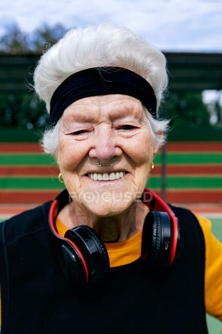 Позитивна зріла жінка з проколотим носом і навушниками в спортивному одязі дивиться на камеру, стоячи на спортивному майданчику під час тренувань — стокове фото