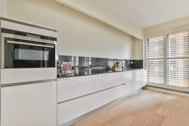 Білі шафи з сучасним обладнанням та різноманітним посудом на стійці в просторій кухні зі стильним інтер'єром у світлій квартирі з вікном — стокове фото