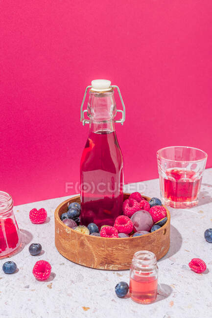 Стеклянная бутылка свежего фруктового сока в деревянной чаше со спелыми ягодами подается на столе со стаканами на розовом фоне — стоковое фото