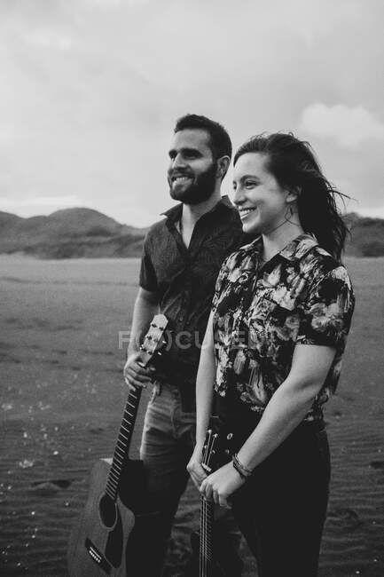Bianco e nero di felice coppia di musicisti in abiti casual in piedi con la chitarra in mano sulla spiaggia di sabbia e guardando lontano di giorno — Foto stock