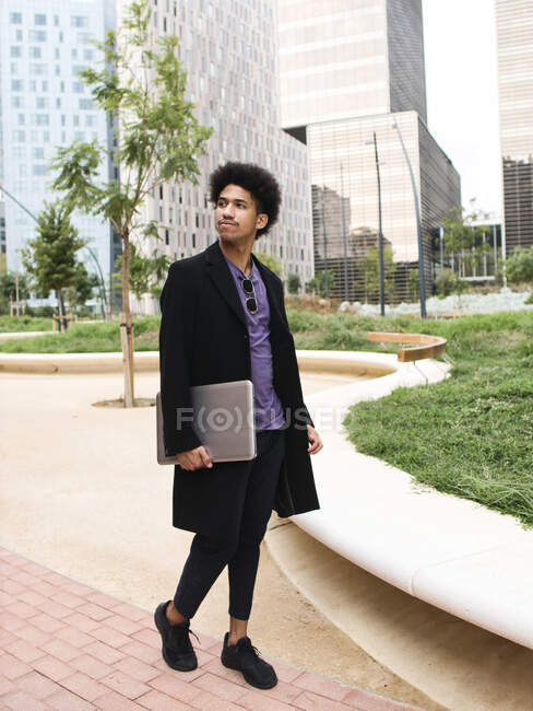 Full body of trendy young ethnic male freelancer with curly dark hair in stylish outfit walking in city park near contemporary skyscrapers with laptop in hand and looking away — Stock Photo