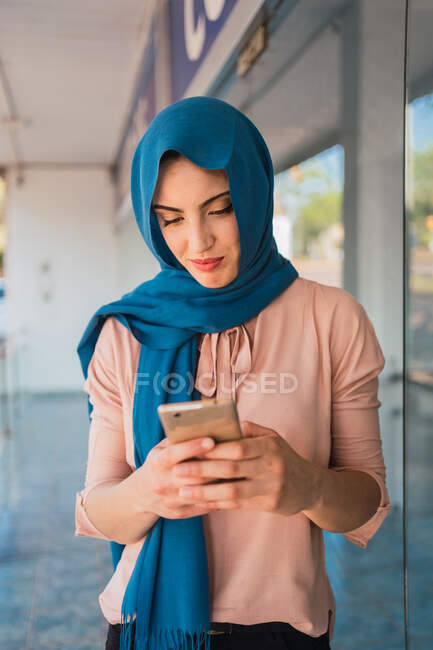 Mujer musulmana en hijab navegando por el teléfono móvil mientras está de pie en la calle de la ciudad - foto de stock